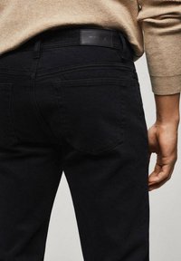 Jean en denim noir à coupe ajustée, avec des poches arrière, un patch logo à la taille, et une texture lisse de couleur foncée.