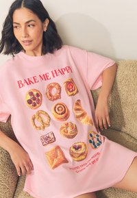 Lichtroze oversized T-shirt met een assortiment aan gebakjesafbeeldingen en rode tekst met de woorden "BAKE ME HAPPY" en "PASTRY OVER PROBLEMS".