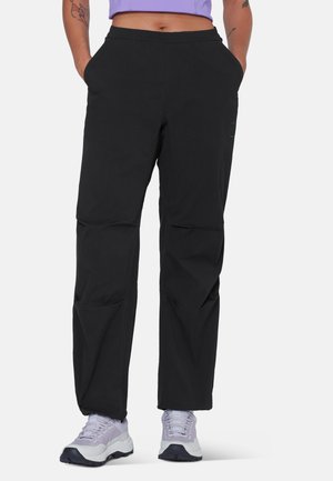 Personne portant un pantalon ample noir avec les mains dans les poches et des baskets violet clair, torse visible de la taille jusqu'aux pieds, fond uni.