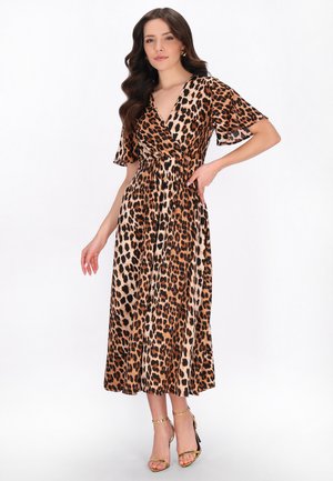 Donna con lunghi capelli mossi che indossa un vestito avvolgente a stampa leopardata con maniche svasate e sandali dorati con tacco alto, posa davanti a uno sfondo bianco.