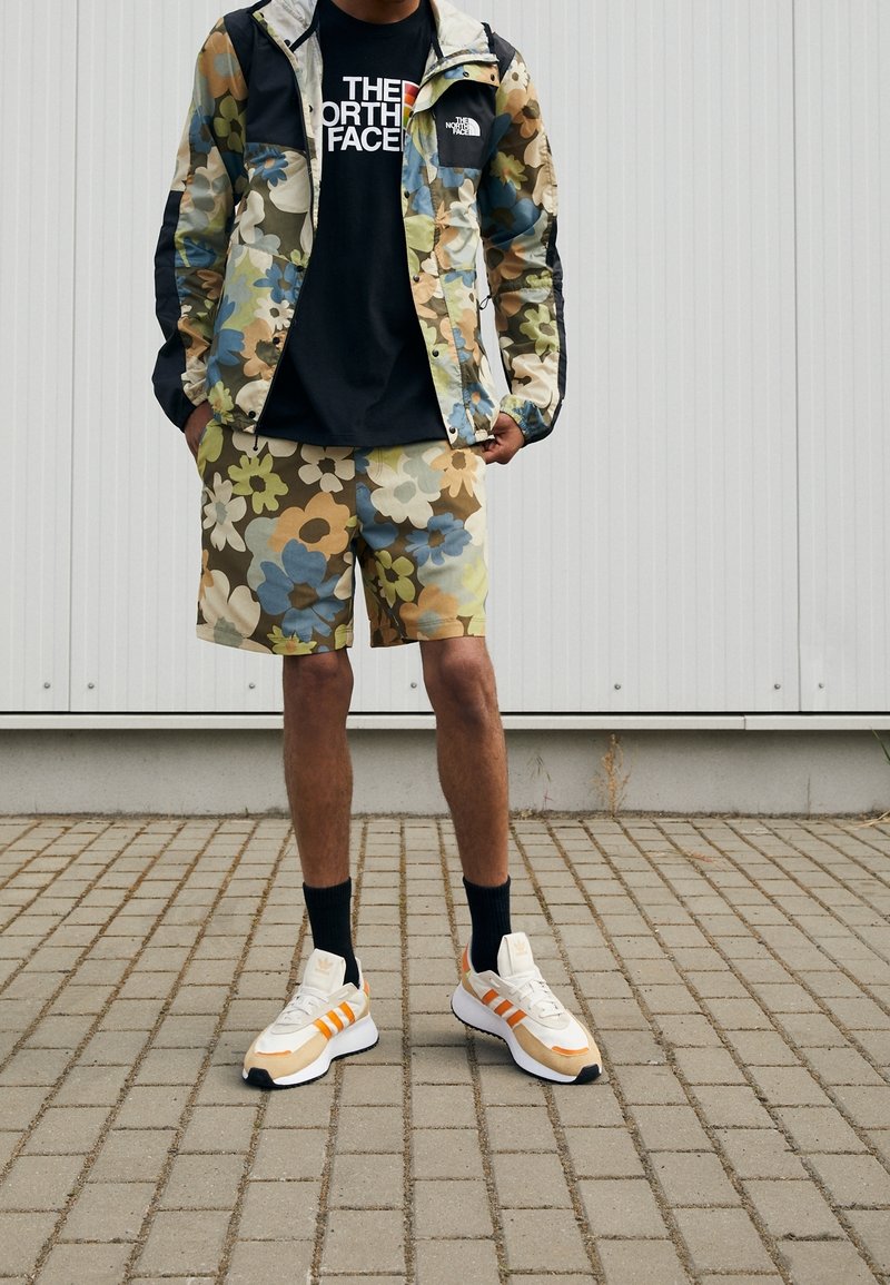Giacca e pantaloni corti con motivo floreale colorato, maglietta nera con design grafico, abbinati a sneakers bianche e arancioni; contesto all'aperto.