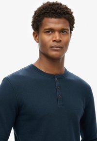 Navy-blaues Langarm-Henley-Shirt aus weichem Stoff, mit Rundhalsausschnitt und drei Holzknöpfen an der Vorderseite.