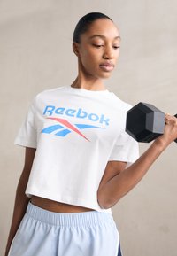 Balta trumpa marškinėliai su mėlynu ir raudonu Reebok logotipu, derinami su šviesiai mėlynomis sportinėmis šortais ir juodu šešiakampiu hanteliu rankoje.