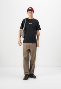 Junger Mann, der ein schwarzes Grafik-T-Shirt, beiger Hose, dunkelbraunen Schuhen, roter Mütze, grauer Schultertasche und einem Tattoo am Unterarm trägt, steht an einer weißen Wand.