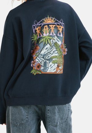 Granatowa bluza z kolorową grafiką gór, palm, kwiatów, słońca i słowem "ROXY" na plecach, noszona z wyblakłymi niebieskimi dżinsami.