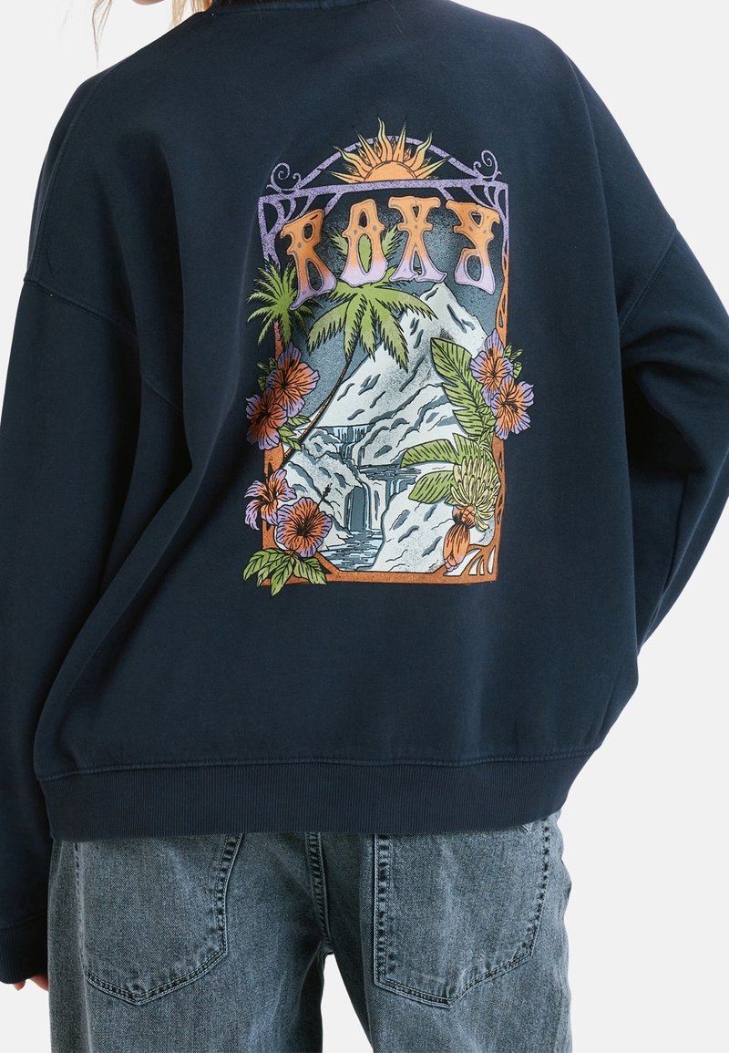 Sweat-shirt bleu marine avec un motif coloré de montagnes, palmiers, fleurs, soleil, et le mot "ROXY" au dos, porté avec un jean bleu délavé.