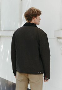 Jachetă maro, texturată, cu guler negru, care are nasturi cu capse la manșete și un croi drept, purtată peste pantaloni bej.
