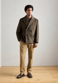 Manteau en laine vert olive à double boutonnage avec quatre boutons noirs, associé à un pantalon en velours côtelé beige et des mocassins marron foncé sur un sol en bois.