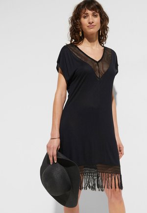 Abito nero con maniche corte, scollo a V caratterizzato da un inserto in rete e orlo frangiato. La modella tiene un cappello nero a tesa larga.