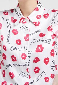 Vit blus med krage, prydd med ett mönster av röda läppar och svart text, tillverkad av ett glänsande material. Knappar synliga fram.