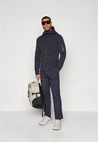 Černá bunda na zip a tmavé úzké kalhoty, se světle šedým a černým batohem. Na sobě má sluneční brýle a bílé tenisky.