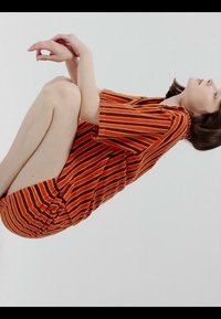 Femme en robe rayée rouge et orange recroquevillée en position fœtale, allongée sur le côté contre un fond blanc uni.