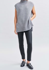 Femme portant un pull gris sans manches à col roulé, un pantalon noir slim et des chaussures noires faciles à enfiler, une main dans la poche.