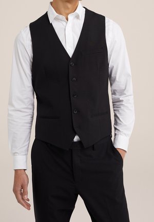 Mann trägt eine schwarze, geknöpfte Weste über einem weißen Langarmhemd und schwarzen Anzughosen, steht mit einer Hand in der Tasche.