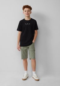 Czarna bawełniana koszulka z białą grafiką, zielone szorty i białe sneakersy na gumowej podeszwie. Model stoi na neutralnym tle.