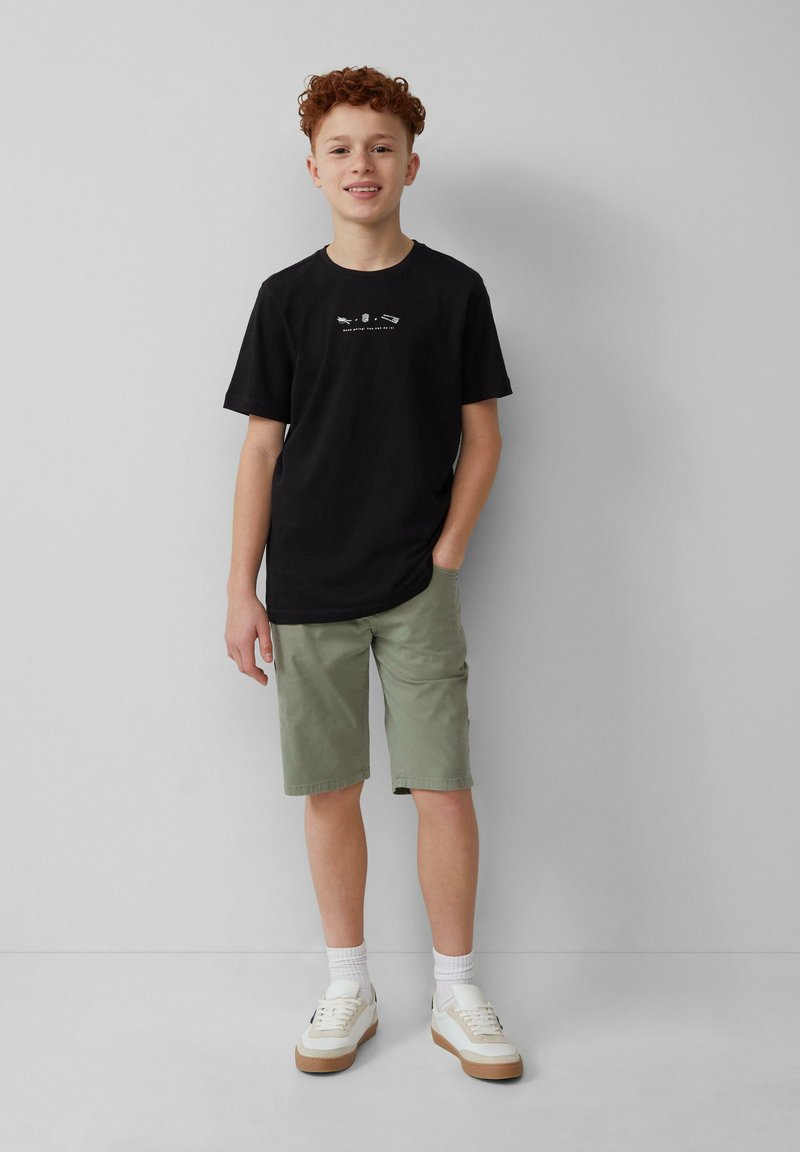 Czarna bawełniana koszulka z białą grafiką, zielone szorty i białe sneakersy na gumowej podeszwie. Model stoi na neutralnym tle.