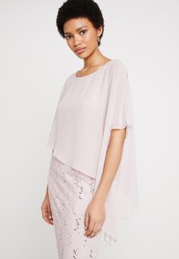 Blouse rose clair avec un design de cape fluide, fabriquée en tissu lisse ; associée à une jupe en dentelle texturée rose avec des sequins.
