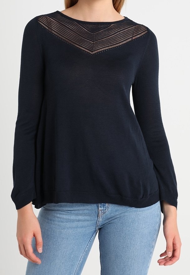 Femme portant un haut à manches longues bleu marine avec un motif en V rayé transparent près du col et un jean bleu clair.