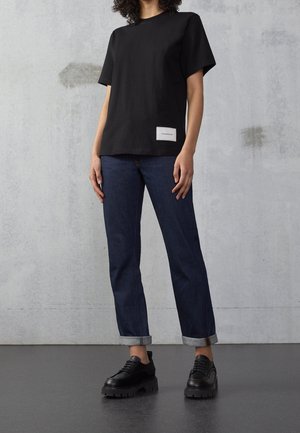Person trägt ein schwarzes kurzärmeliges Shirt mit einem weißen Etikett, dunkelblaue hochgekrempelte Jeans und schwarze klobige Schnürschuhe.
