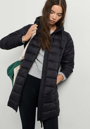 Frau trägt eine lange schwarze Steppjacke mit Kapuze, grauen Sweatshirt, schwarze Hose und trägt eine grün-beige Tragetasche.