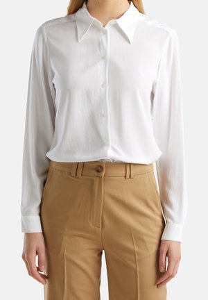 Vrouw draagt een wit, knoopsluitend overhemd met lange mouwen, ingestopt in hooggetailleerde beige broek met knoop en riemlussen.