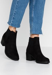 Czarne zamszowe botki za kostkę z falbaniastymi brzegami, masywnym obcasem i teksturowaną cholewką. Noszone z przyciętymi niebieskimi dżinsami.