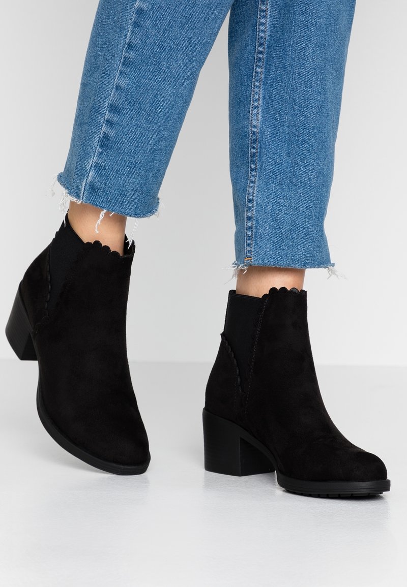 Czarne zamszowe botki za kostkę z falbaniastymi brzegami, masywnym obcasem i teksturowaną cholewką. Noszone z przyciętymi niebieskimi dżinsami.