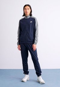 Person standing wearing a navy and gray Nike tracksuit with white sneakers on a blue floor and white background.