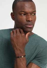 Svart perlebånd med gullaccent i minimalistisk design. Båret på et lyst håndledd. Modellen har kort hår og har på seg en grønn skjorte.