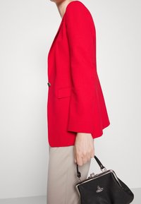 Červený strukturovaný blazer s dlouhými rukávy, s jedním knoflíkem, v kombinaci s černou kabelkou s kovovým zapínáním a krátkým uchem.