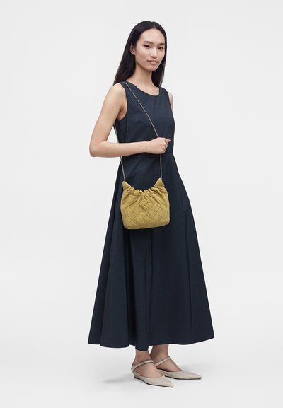 Femme en robe noire sans manches tenant un sac à main matelassé moutarde avec une bandoulière chaîne, chaussée de souliers beiges à talons bas.