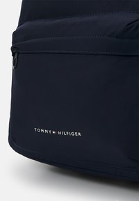 Zaino in tessuto blu navy con una tasca frontale con zip, caratterizzato da cuciture a contrasto e un logo bianco: "TOMMY HILFIGER" nella parte inferiore.