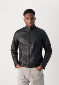 Jachetă din piele neagră cu guler înalt, textură matlasată pe umeri, buzunare frontale și închidere cu fermoar. Purtați peste blugi de culoare deschisă.