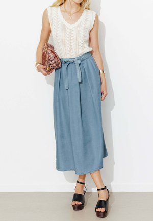 Femme portant un top sans manches en maille blanche et une jupe bleue taille haute avec ceinture nouée, tenant une pochette à motifs et portant des sandales compensées noires.