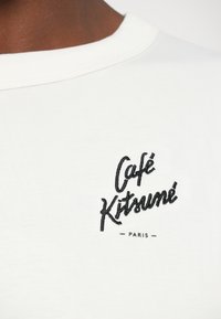 T-shirt bianco in cotone con scollo rotondo e texture a costine, caratterizzato da un ricamo nero con il marchio: "Café Kitsuné Paris" al centro.