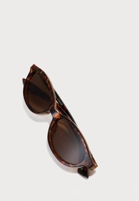 Sonnenbrille aus Schildpatt mit dunkelbraunen Gläsern, runder Form, dicken Rahmen und glänzender Oberfläche. Mit spitzen Enden und subtiler Markenkennzeichnung an der Seite.