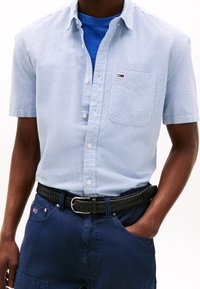 Košile s krátkým rukávem v světle modré kostce s bílými akcenty, límečkem na knoflíky a přední kapsou, nošená přes modré tričko a tmavě modré kalhoty.