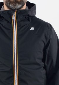 Čierna nepremokavá bunda s kapucňou, kontrastným žltým, červeným a čiernym pruhom pozdĺž zipsu a bielym logom na hornej ľavej strane.