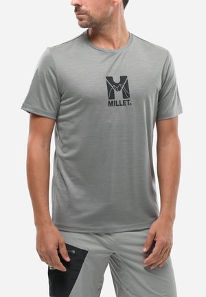 Homme portant un t-shirt à manches courtes gris clair avec le logo "MILLET" noir et un pantalon gris clair avec une poche zippée noire.