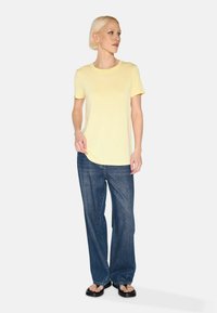 Lichtgele t-shirt met korte mouwen, relaxte pasvorm, gecombineerd met losse blauwe jeans. Model staat in een neutrale omgeving, draagt zwarte sandalen.
