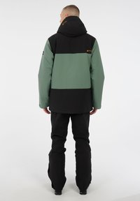 Giacca isolante verde e nera con cappuccio, caratterizzata da un design bicolore, dettagli regolabili e un logo sulla spalla sinistra.