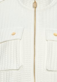 Veste en tissu blanc texturé avec fermeture éclair dorée et deux poches avant à rabat sécurisées par des boutons ronds dorés.