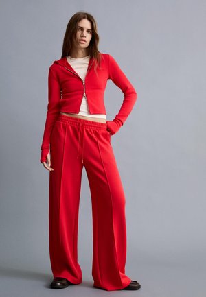 Woman standing in red zip-up hoodie and wide-leg red pants with beige top and dark shoes against gray background.