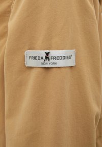 Tan tyg som har en sydd etikett med "FRIEDA FREDDIES" och en hundlogotyp. Etiketten är vit med svart text och detaljer.