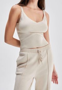 Žena v béžovém žebrovaném pleteném cropped topu se špagetovými ramínky a odpovídajících kalhotách s vysokým pasem na šňůrku, stojící na jednobarevném pozadí.