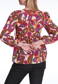 Kleurige blouse met een paisleypatroon in rood, groen en paars, met pofmouwen en een geaccentueerde taille, gecombineerd met donkere jeans.