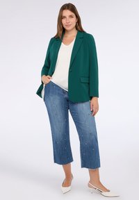 Femeie îmbrăcată cu un sacou verde, bluză albă cu decolteu în V, blugi scurți albaștri cu embellishments și tocuri albe ascuțite, stând pe un fundal alb.