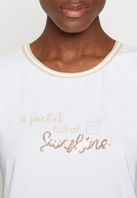 Hvid t-shirt med guld-accenteret halsudskæring og skinnende guldtekst "a pocket full of Sunshine", der indeholder en lille lommegrafik i designet.