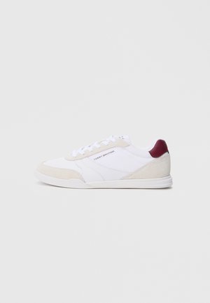 Witte lage sneaker met beige suède accenten, een maroonkleurige hielpatch, witte veters en het "Tommy Hilfiger"-logo aan de zijkant.