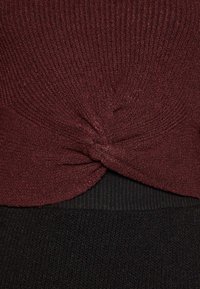 Bordeauxkleurige top met een textuur van ribbelstof. Heeft een geknoopt detail bij de taille en is gecombineerd met een zwarte gebreide onderkant.